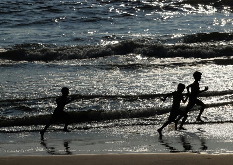 Three boys running along a beach in India at sunset, silhouetted against waves.